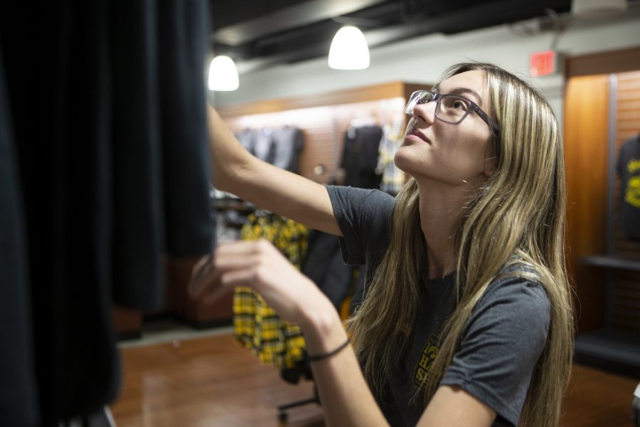 Iowa Hawk Shop employee Medeline Recker straightens a shirt hanging on a rack on Thursday, Oct. 31, 2019. Recker, who has been working at the shop located in the basement of the IMU since January, is a sophomore studying art with a focus on graphic design. She said most of her time on the clock is spent tidying up the merchandise and folding clothes. 